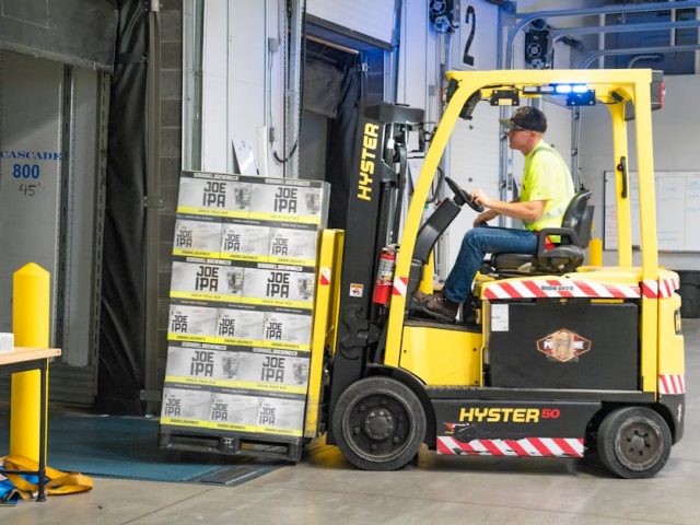 A worker drives a Hyster forklift moving Joe IPA boxes in a warehouse.